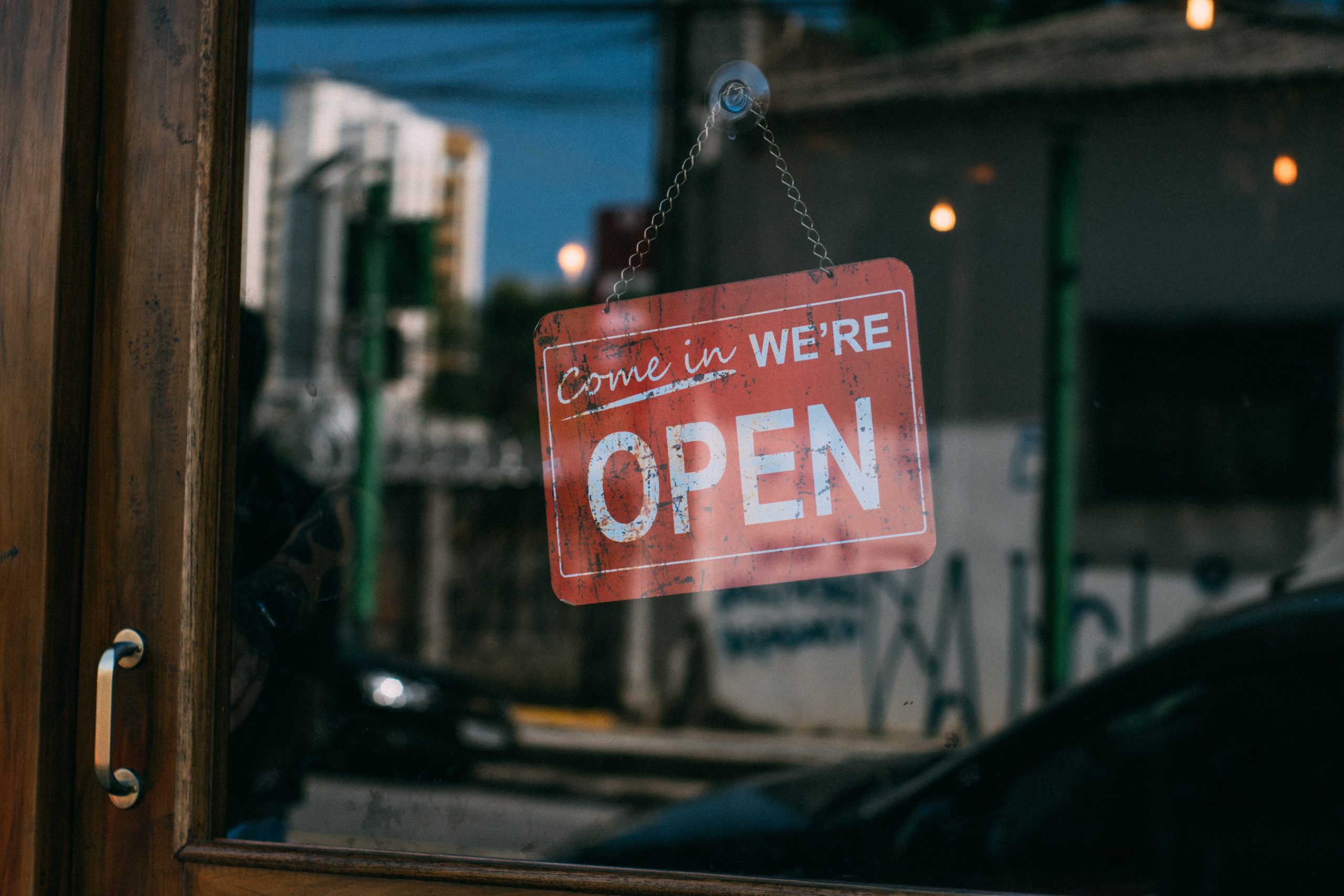 red sign that reads "come in we're open" in white text, hanging from a glass door