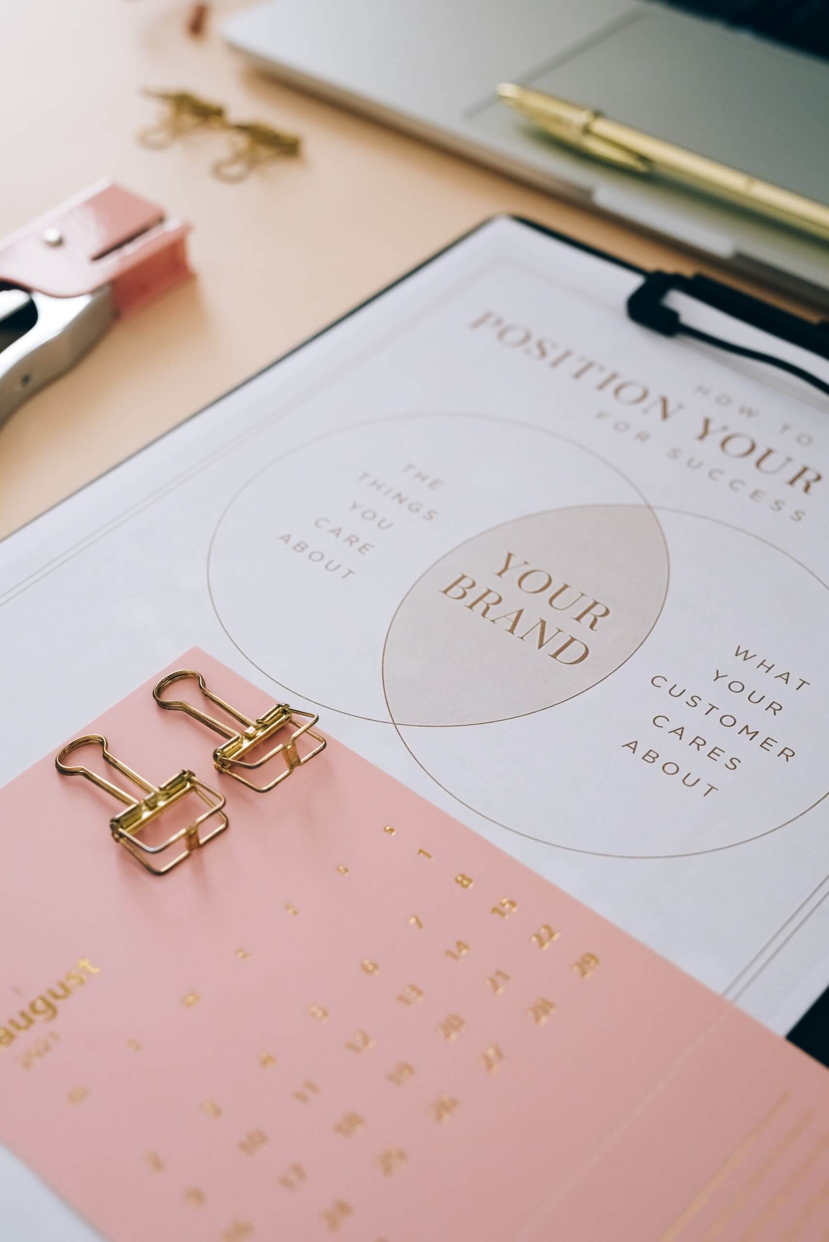 table with pink calendar, stapler, laptop, and and clipboard that reads "Your Brand"